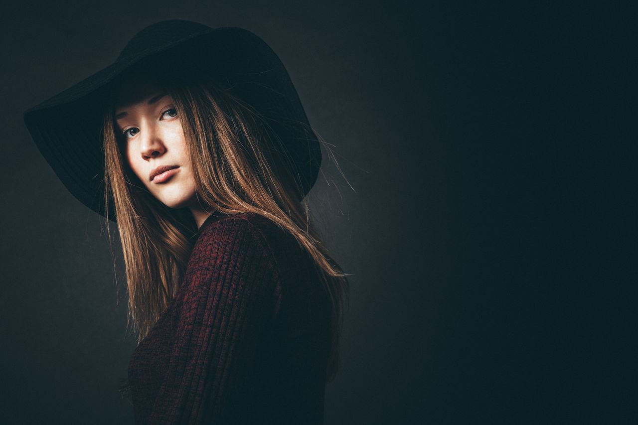 services-02 Studio portrait of a young woman wearing a black hat against a dark background, showcasing elegance and style.