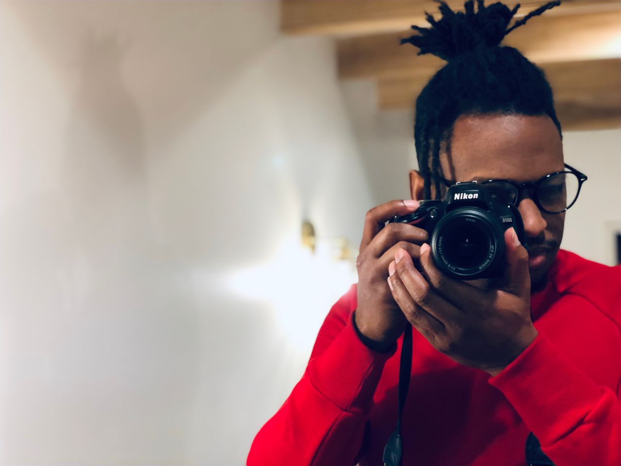 Crafting Captivating Headlines: Your awesome post title goes here A man in red sweater holds a professional camera indoors, capturing a self-portrait.