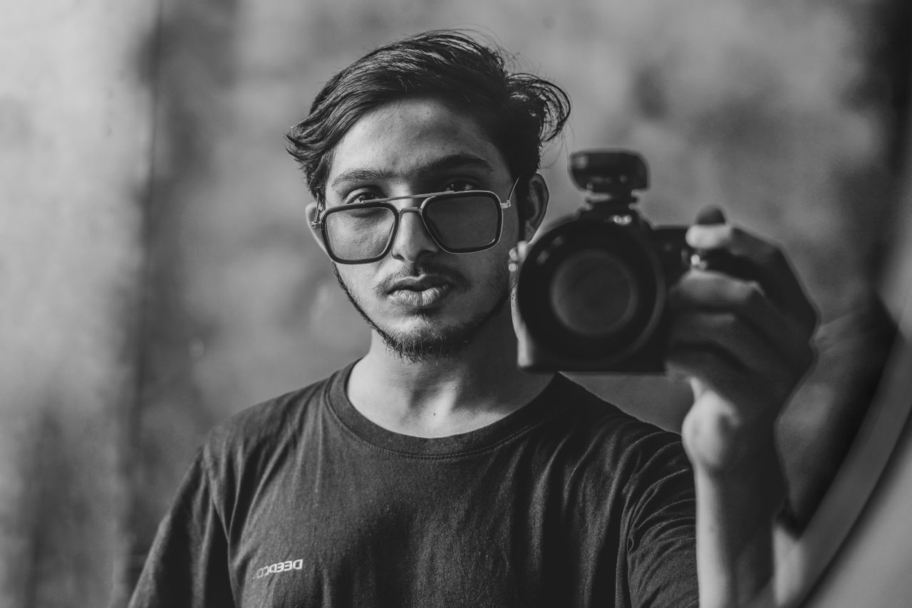 services-01 A stylish black and white self-portrait of a man holding a camera, wearing sunglasses.